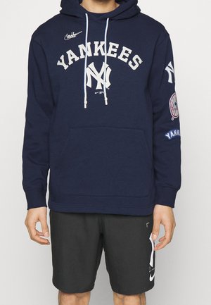 Personne portant un sweat à capuche Yankees bleu marine avec des lettres blanches et un short noir avec un logo Nike blanc, debout devant un fond uni.