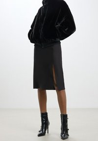 Černá bombera z umělé kožešiny s žebrovanými manžetami, doplněná černou pouzdrovou sukní ke kolenům s bočním rozparkem. Dokončeno černými kotníkovými botami.