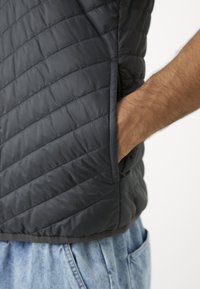 Black quilted vest featuring a smooth, lightweight material, side pockets, and a sleek design, worn over light blue jeans.