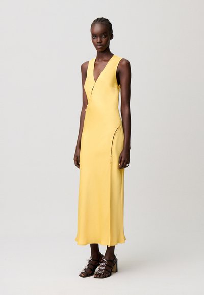 Donna dalla pelle scura e capelli intrecciati che indossa un vestito midi giallo senza maniche con bottoni laterali e sandali neri con tacco su sfondo semplice.