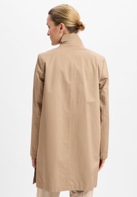 Beige lange jas met hoge kraag, gemaakt van soepel materiaal. Heeft een rechte silhouet en zijsplitsen voor bewegingsvrijheid.