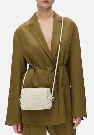 Sac à bandoulière tissé beige avec un motif texturé et une sangle blanche. Porté avec un blazer vert olive doté d'une ceinture à nouer et de détails de poches.