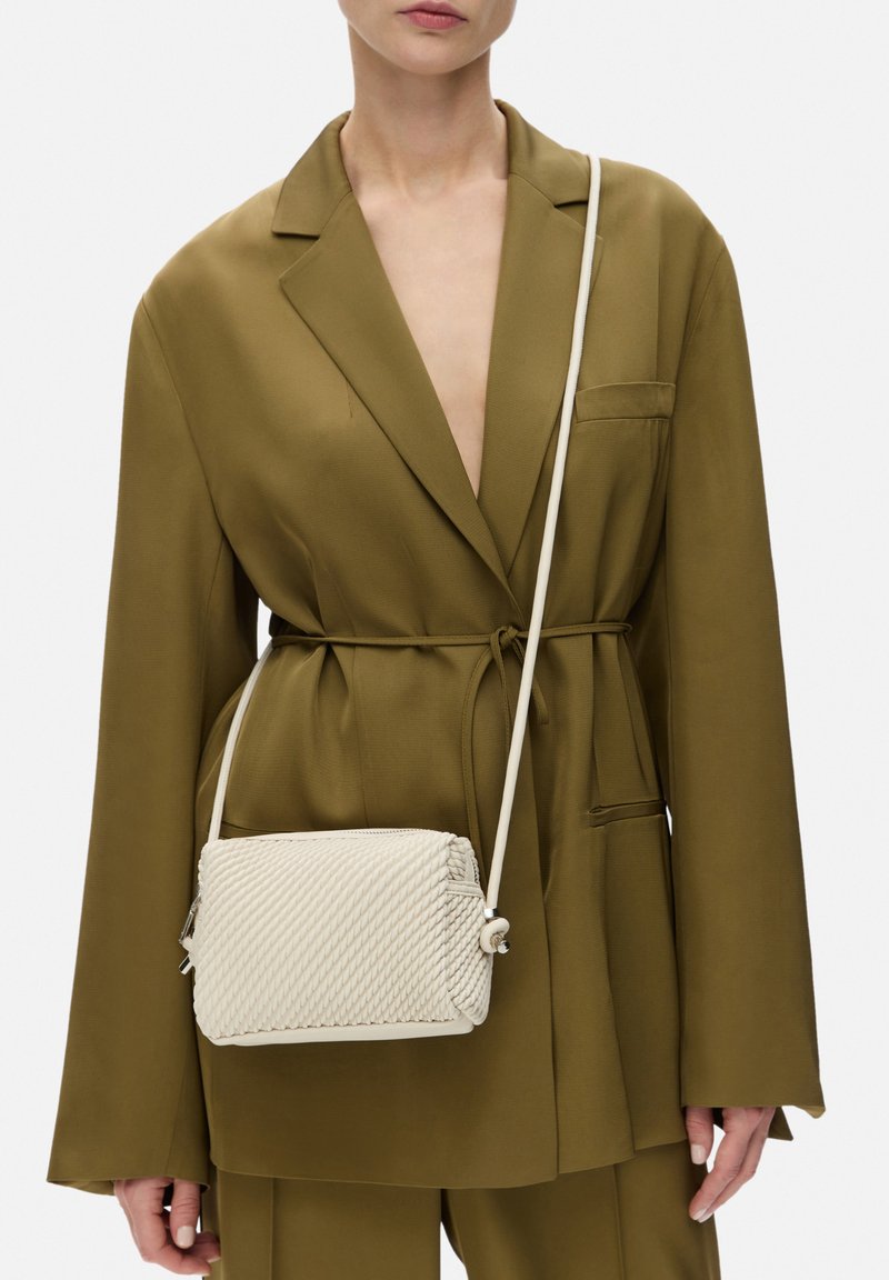 Bolso cruzado tejido beige con un patrón texturizado y tira blanca. Se lleva con un blazer verde oliva que presenta un lazo en la cintura y detalles en los bolsillos.