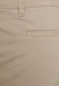 Tessuto di cotone beige con una texture a coste, dotato di una piccola tasca posteriore e dettagli cuciti lungo la cintura e le cuciture.
