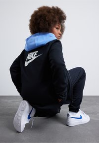 Mustan tõmblukkidega jakiga, millel on sinine kapuuts, ning seljal suur valge Nike logo. Valged tossud sinise Nike logo kaubamärgiga. Tekstuuriga juuksed.