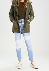 Olive green parka with a fur-trimmed hood, paired with light blue patchwork jeans and white sneakers with beige and orange accents.
