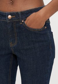 Vaqueros de mezclilla azul oscuro con un diseño ajustado, herrajes de cobre, costuras visibles y bolsillos laterales. La tela tiene una textura suave.