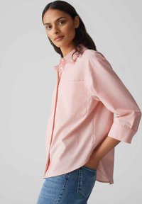 Helles, rosa Button-up-Hemd mit einer einzelnen Brusttasche, hochgekrempelten Dreiviertelärmeln und einem lässigen Schnitt, kombiniert mit blauen Jeans.