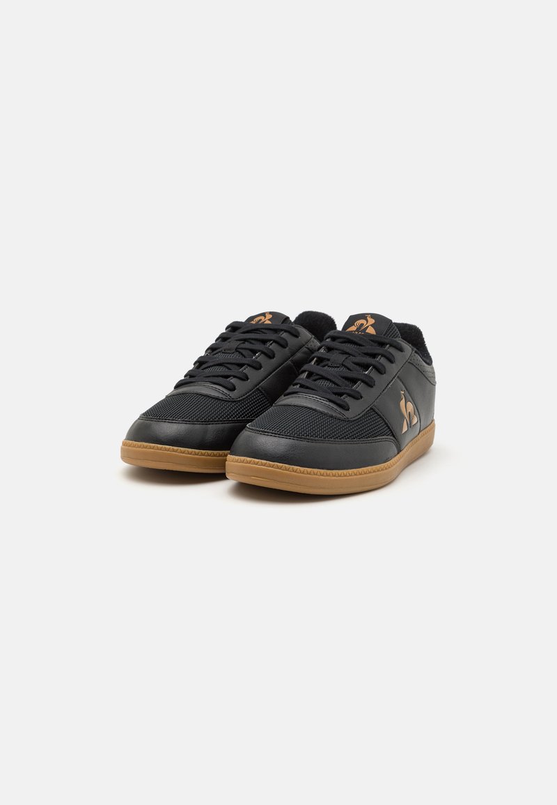 Zapatillas deportivas negras con parte superior de malla y cuero, suela de goma y cordones a tono; cuenta con un pequeño logo dorado en el lado.