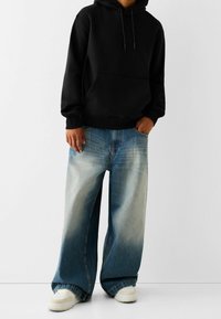 Persona vestida con una sudadera con capucha negra, jeans anchos de mezclilla azul deslavado y zapatillas blancas, de pie con una mano en el bolsillo frente a un fondo liso.