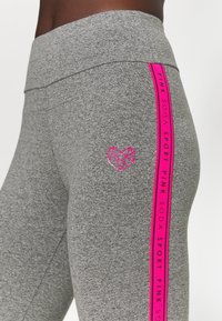 Szare legginsy sportowe z różowym bocznym paskiem z napisem "PINK SODA SPORT." Obecne jest małe różowe logo w kształcie geometrycznego serca.