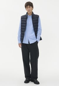 Marinblå vadderad väst över en ljusblå skjorta med knappar, kombinerad med svarta byxor och svarta snörskor. Enkel, strukturerad design.