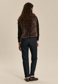 Vrouw met lang krullend haar, gekleed in een leopardprint bontvest, een zwarte langesliekenshirt, donkere opgerolde jeans en bruine loafers, staande met haar rug naar de kijker gericht.