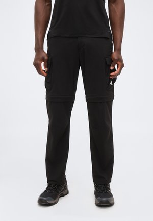 Mann trägt schwarze wandelbare Cargohosen mit abnehmbaren Shorts, schwarze Sneaker und schwarzes Hemd, steht vor einem schlichten hellen Hintergrund.