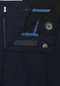 Sötétkék, sima tapintású anyag, gombbal és kontrasztos kék varrással. Cipzáras zsebbel és egy kis ezüst kiegészítővel rendelkezik.
