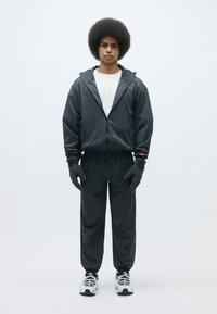 Jeune homme avec une coupe afro portant une veste à capuche noire, un pantalon noir, des gants gris et des baskets blanches, se tenant contre un fond blanc.