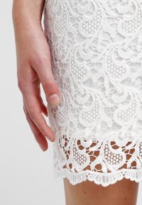 Main reposant contre un tissu en dentelle blanche avec un motif floral et géométrique sur un vêtement.