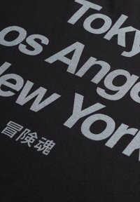 Sort t-shirt med stor, fed, hvid tekst, der navngiver byer: Tokyo, Los Angeles, New York, med ekstra tekst i en mindre skrifttype.