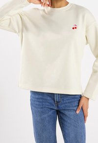 Persona che indossa una camicia a maniche lunghe color crema con un piccolo ricamo di ciliegia rossa sul petto e jeans in denim blu, con la mano appoggiata sull'anca.