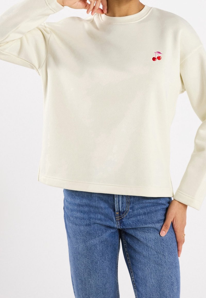 Persona che indossa una camicia a maniche lunghe color crema con un piccolo ricamo di ciliegia rossa sul petto e jeans in denim blu, con la mano appoggiata sull'anca.