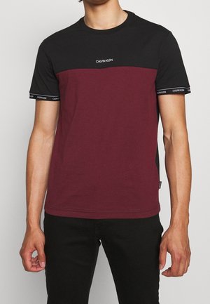 Muž nosící tričko s krátkými rukávy a kulatým výstřihem, s černým vrškem, vínově červeným tělem a bílým logem "Calvin Klein" na hrudi a rukávech.