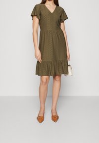 Oliivinvihreä polvipituinen mekko, jossa on V-aukko, lyhyet röyhelöistetyt hihat, kuvioitu kangas ja levenevä helma. Yhdistetty vaaleanruskeisiin kärjellisiin ballerinoihin.