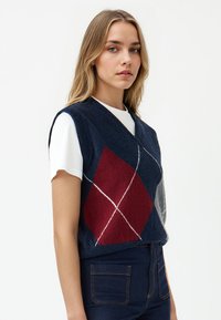 Gilet lavorato a maglia senza maniche di colore blu navy con scollo a V, caratterizzato da motivi argyle rossi, grigi e bianchi. Indossato sopra una maglietta bianca, parte di jeans scuri.