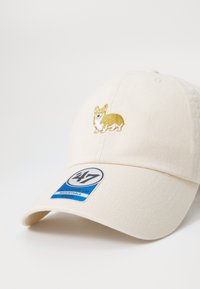 Krém színű baseball sapka, melyen egy arany hímzésű corgi található. Állítható pánttal és egy márkajelzéssel rendelkezik az oldalon. Texturált anyagból készült.