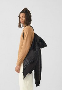 Een jonge man met dreadlocks staat zijwaarts en draagt een tan kleurig shirt met lange mouwen, witte broek en een zwarte gewatteerde jas over één schouder.