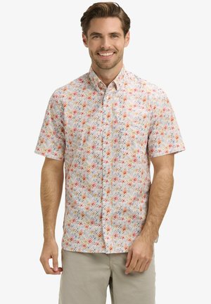 Homme souriant portant une chemise à manches courtes boutonnée avec de petits motifs floraux rouges, orange et violets, et un pantalon beige clair.