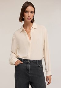 Blusa color crema con bottoni sul davanti, maniche lunghe e polsini arricciati, abbinata a pantaloni in denim scuro con cuciture decorative.