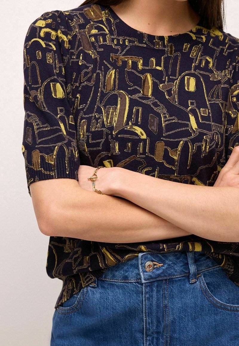 Femme portant un haut noir à manches courtes avec un motif abstrait de bâtiment jaune et marron, un jean bleu, et un bracelet chaîne en or, bras croisés.
