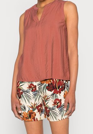 Bezrekawka bluzka w kolorze rdzy połączona z białymi szortami ozdobionymi dużymi czerwonymi, pomarańczowymi kwiatami hibiskusa i zielonymi liśćmi palmowymi.