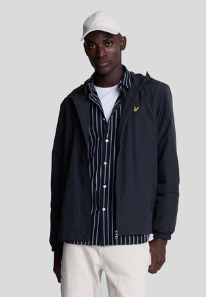 Schwarzer Mann mit weißer Kappe, marineblau gestreiftem Hemd, schwarzer Jacke mit gelbem Logo und weißer Hose vor hellgrauem Hintergrund.