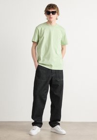 Light green cotton T-shirt, dark denim wide-leg pants, and white sneakers. Model wearing black rectangular sunglasses. Plain background.