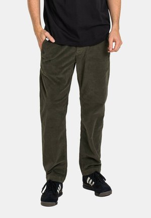 Man wearing olive green corduroy pants, black sneakers with white stripes, and a black t-shirt, standing with one hand in pocket.