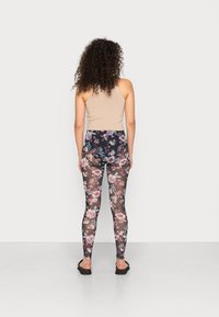 Leggings florais com uma textura preta transparente e padrões de flores roxas, rosas e azuis, combinados com uma blusa ajustada bege e sandálias.