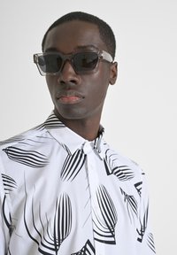Camisa blanca de manga corta con estampado de hojas negras; combinada con gafas de sol de armazón transparente anguloso con lentes oscuros.