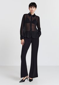 Camicia nera trasparente con bottoni, maniche lunghe, abbinata a pantaloni neri a vita alta e décolleté nere con punta.
