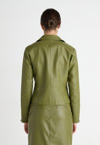 Veste en cuir vert au design ajusté, avec un col en pointe, des coutures verticales et des accents en matériel argenté. Texture lisse.