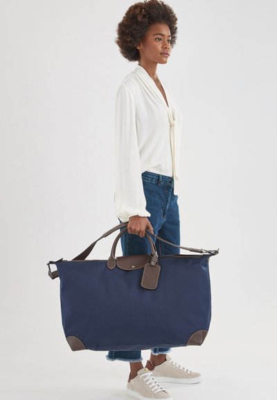 Marineblaue Duffel-Tasche mit braunen Lederakzenten und Traggriffen, ausgestattet mit einem Gepäckanhänger. Hergestellt aus robustem Stoff, geräumiges Design.
