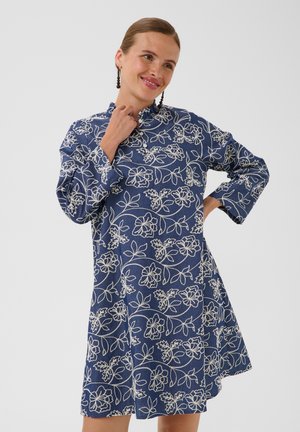 Femme souriante portant une robe bleue avec une broderie florale blanche, manches longues et col montant, debout devant un fond clair uni.