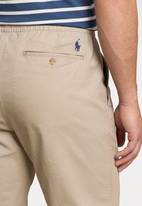 Beiget housut, joissa on joustava vyötärönauha, napillinen takatasku ja pieni sininen polo-logo takana. Sileä kangas ja istutettu malli.