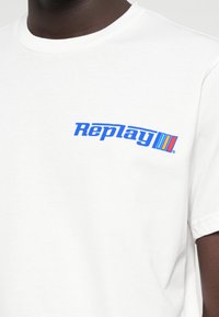 Λευκό βαμβακερό μπλουζάκι με μπλε λογότυπο "Replay" στην αριστερή πλευρά του στήθους. Κοντά μανίκια με σχέδιο λαιμού τύπου crew. Λείο υφή.
