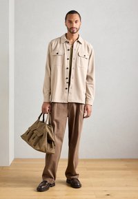 Bărbat îmbrăcat cu o jachetă bej cu butoni și pantaloni maro, ținând o geantă mare din piele maro, stând pe un podea din lemn deschis împotriva unui perete simplu.