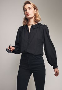 Črna bluzica s puhovimi rokavi in naboranim ovratnikom, v kombinaciji s temnimi skinny džinsami. Model drži črne sončne očala.