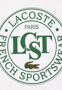T-shirt blanc avec un logo circulaire vert comportant les inscriptions "LACOSTE", "PARIS", "LCST" et "FRENCH SPORTSWEAR", ainsi qu'un crocodile vert brodé.