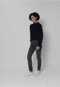 Svart polotröja, figurnära mörka jeans och vita sneakers. Lockigt hår, minimalistisk design mot en ljusgrå bakgrund.