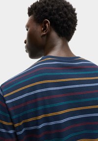 Uomo con capelli ricci corti di profilo, che indossa una camicia blu navy con strisce orizzontali rosse, gialle, verdi e azzurro chiaro.