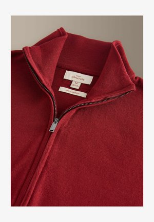 Rød merino uld trøje med lynlås og høj krave. Har en sølv lynlås og mærkater, der angiver størrelse og materiale.